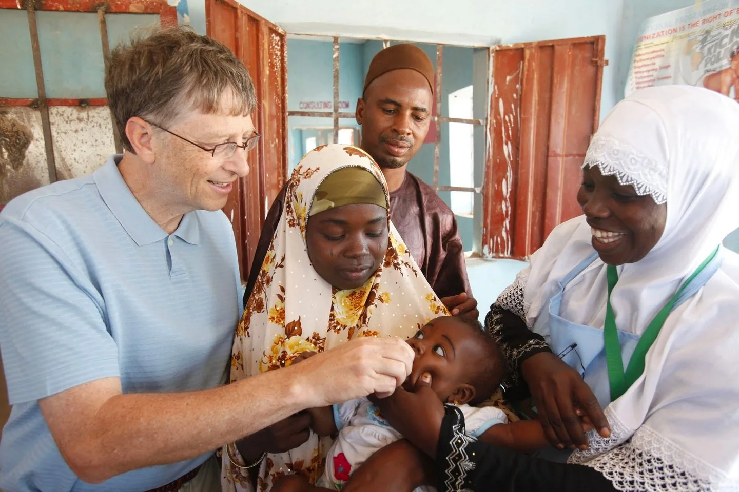 Bill Gates is depicted in the image, participating in a healthcare activity. He is smiling and appears engaged, wearing a light blue polo shirt, and glasses. He is assisting with administering an oral vaccine to a young child held by a woman wearing a hijab with a floral pattern. Another woman, dressed in a white hijab and apron, is also present, smiling warmly, possibly a healthcare worker. A man in a brown outfit and cap stands behind them, observing the interaction. The setting is indoors, likely in a clinic or healthcare facility, with blue walls and open windows in the background. There is a sense of warmth and collaboration in the scene, highlighting a moment of community health support and philanthropy.