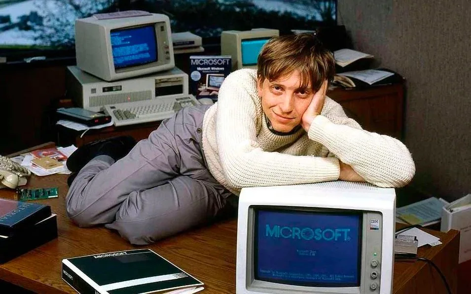 This image features Bill Gates posing in a vintage office setting. He is casually lying on a desk, resting his head on his hand, and smiling at the camera. Gates is wearing a light-colored sweater and gray pants, contributing to a relaxed and approachable demeanor. The office is filled with computer equipment and documents, emphasizing a technological theme.
In the foreground, an IBM monitor prominently displays the word "MICROSOFT," highlighting the brand's significance. Behind Gates, another computer setup and various office supplies are visible, reinforcing the professional yet informal atmosphere. The lighting is natural, likely from a window, which creates a warm and inviting ambiance. The image encapsulates a moment from the early days of personal computing, capturing both the person and the pioneering spirit of the era.
- Title: "Microsoft Windows"