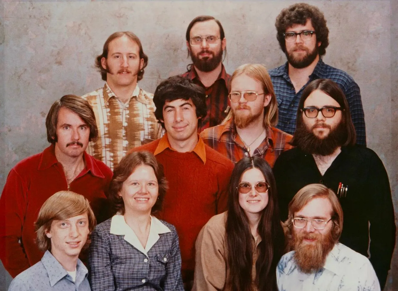 This image is a vintage group photo featuring Bill Gates and several others from the early days of Microsoft. The group is posed against a simple, textured backdrop typical of the 1970s. There are twelve people in total, arranged in three rows. Bill Gates, identified through the hint, is in the front row on the left, smiling slightly, with distinctive longer hair and wearing a light blue shirt underneath a sweater.
The mood of the photograph is casual yet formal, with most of the individuals exhibiting neutral or slight smiles. The attire is distinctly reflective of the 1970s, with patterned shirts and a mix of sweaters and collared shirts. A few of the men have notable facial hair, such as mustaches and beards, contributing to the period style. The lighting is even, suggesting a studio setup, emphasizing the features and attire of the group members. This image captures an important moment in technology history, likely representing an early team at Microsoft.