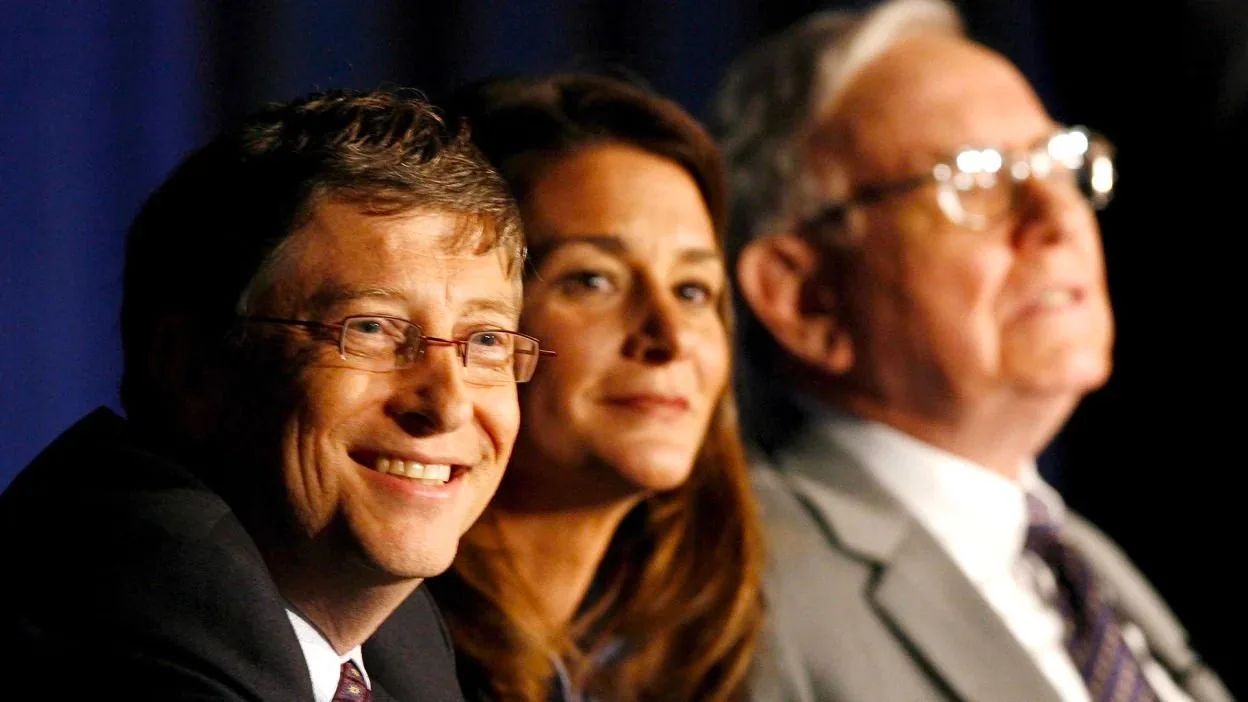 The image depicts a panel setting at a conference or event, featuring three individuals seated together. The focus is on Bill Gates, who is smiling and wearing glasses. He is dressed in a formal suit with a white shirt and a patterned tie, suggesting a professional occasion. To his right, there is a woman with long brown hair, and to her right, an older man wearing glasses and a light-colored suit. The lighting is warm, highlighting their faces against a darker background, which adds to the formal and serious atmosphere of the event. The expressions suggest a moment of engagement or anticipation, possibly during a discussion or speech. The photograph captures a candid moment, emphasizing their attentive and positive demeanor.