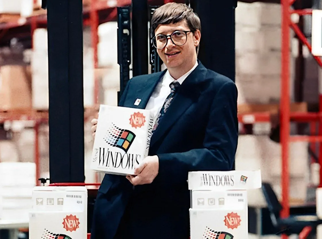 Bill Gates is depicted in this image holding a box of Microsoft Windows. The setting appears to be indoors, possibly a warehouse, as suggested by the industrial shelving and boxed inventory in the background. Gates is dressed in a dark suit with a tie, and he has a cheerful expression, suggesting a promotional context. The Windows boxes prominently feature the classic Windows logo with a colorful flag-like icon and the word "NEW" highlighted, indicating a product launch or announcement. The lighting is even and natural, contributing to a professional yet casual atmosphere. The image likely dates back to when a new version of Windows was being introduced, capturing a moment in tech history. The overall tone is positive and business-oriented, reflecting Gates's role in the software industry.