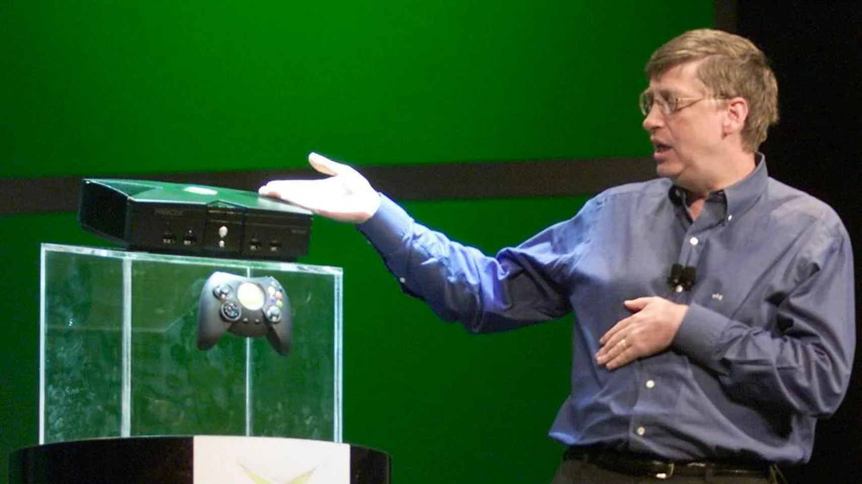 Bill Gates is presenting the original Xbox console. He stands beside a glass podium with the Xbox and its controller on display. Gates is wearing a blue button-up shirt and glasses, gesturing towards the console with a slight smile. The setting suggests a formal presentation, likely at a technology event. The Xbox, black and rectangular, is elevated for visibility, while its controller hangs below. The backdrop is a vivid green, emphasizing the Xbox branding. The lighting is bright, focusing on both Gates and the console, highlighting the significance of the momentous product launch. This scene captures a key moment in the gaming industry, marking Microsoft's entry into the console market.