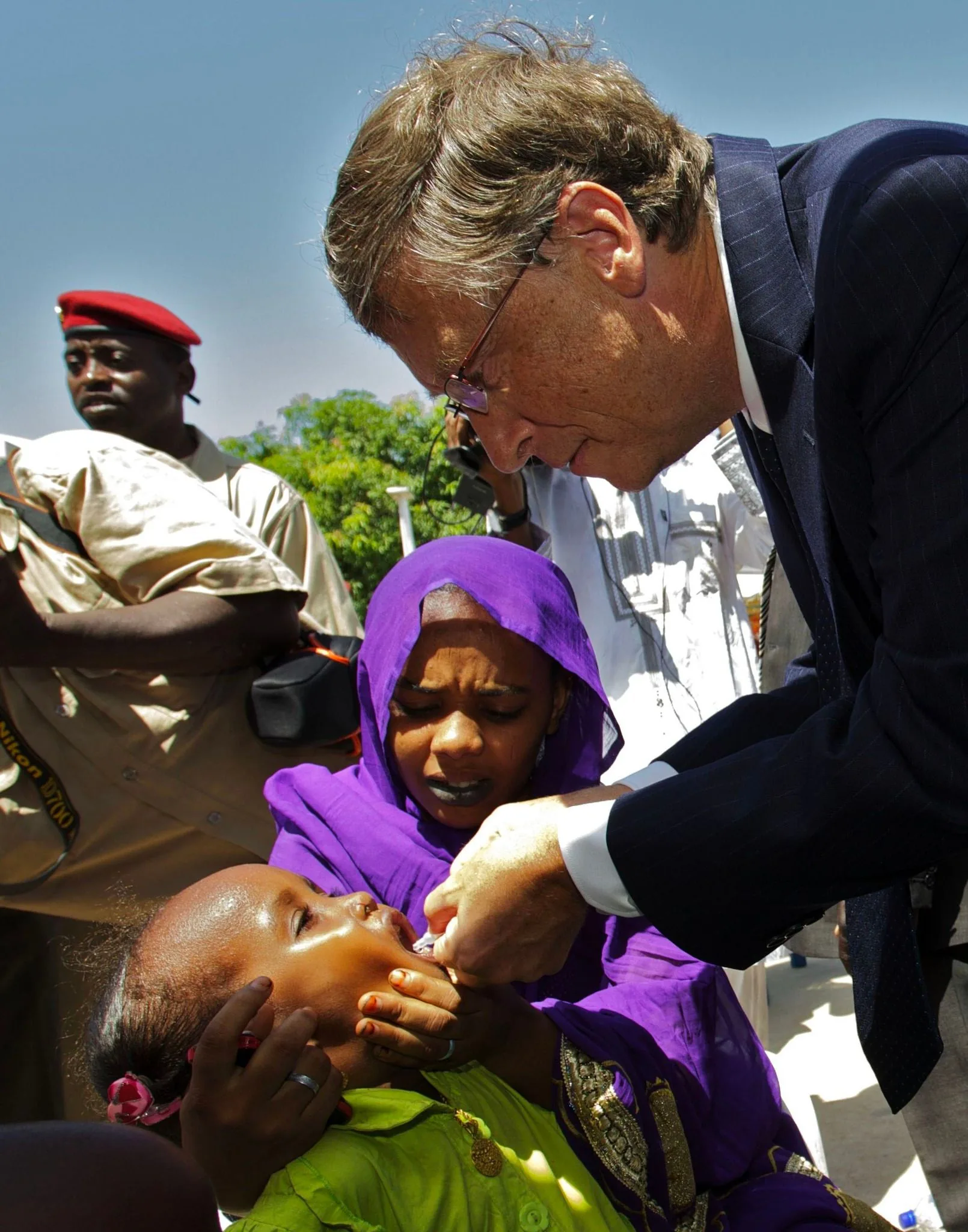 The image captures a moment where Bill Gates is administering an oral vaccine to a young child. He is wearing a dark suit and glasses, leaning forward with a focused and gentle expression as he carefully drops the vaccine into the child's mouth. The child, dressed in a vibrant green shirt, is held by an older girl who is wearing a purple hijab, showing a mix of apprehension and care. The setting appears to be outdoors, likely in a community or healthcare setting. Behind Bill Gates, there are other individuals present, including a man in a uniform with a red beret, suggesting a location with organized healthcare assistance. The lighting is bright and natural, indicating a sunny day. This scene highlights a philanthropic effort focused on vaccination and global health.
