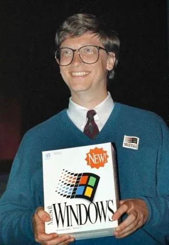 Bill Gates is depicted in this image, smiling at a launch event. He is holding a boxed copy of Microsoft Windows, prominently displaying the Windows logo and the word "NEW" on the packaging. Gates is dressed in a blue sweater over a white shirt, complemented by a patterned tie. He also wears large glasses, indicative of the fashion of the time. A small button with the Windows logo is pinned to his sweater, further emphasizing the event's focus on this software release. The background is dark, making the subject and the box stand out more vividly. The overall mood is one of pride and accomplishment, typical of a product launch setting. The image captures a significant moment in technology history, highlighting the introduction of a major software product.