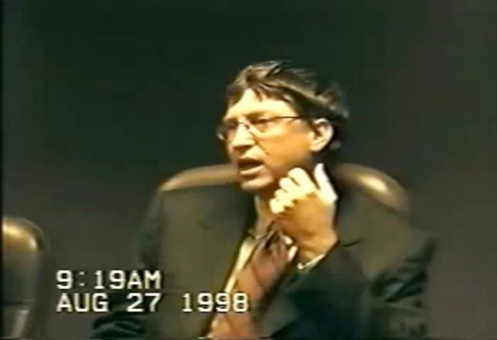 Bill Gates is captured in an indoor setting, dressed formally in a dark suit and tie, indicative of a professional environment, likely a legal deposition. His posture is slightly reclined in a chair, with one hand raised, as if making a point or responding to a question. His expression appears serious and engaged, typical of a formal interview or deposition scenario. The lighting is subdued, and the background is plain, focusing attention on Gates himself. On-screen, there is a timestamp reading "9:19AM AUG 27 1998," suggesting the date and time of the recording, contributing to the formal and archival nature of the scene. The overall tone is serious, reflecting a moment of legal or corporate significance.