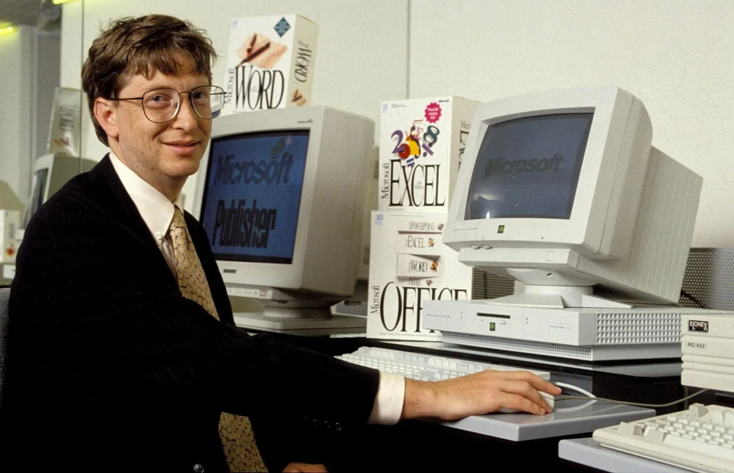 Bill Gates is shown seated at a desk in an office setting, surrounded by computer equipment and software boxes. He is wearing a dark suit with a white shirt and a patterned tie, looking directly at the camera with a slight smile. In front of him is a CRT monitor displaying "Microsoft Publisher" on the screen. Nearby, another monitor shows the word "Microsoft." Stacked beside the monitors are boxes of software including "Microsoft Word," "Microsoft Excel," and "Microsoft Office," indicating the focus on Microsoft's software products. The scene is brightly lit, highlighting the technological theme. Bill Gates’ relaxed posture and expression suggest a confident and promotional context. The equipment, such as the monitors and keyboards, reflect the early computing era, reinforcing the historical significance of the image.