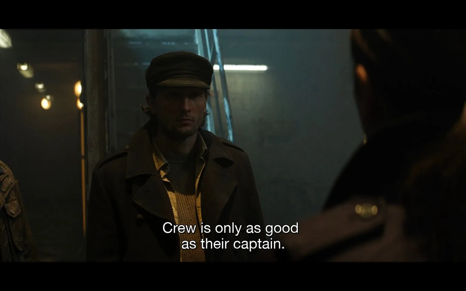 ```
**SECTION 2**
The image shows a dimly lit indoor scene with a man standing in the foreground, partially obscured by shadow. The atmosphere is tense and dramatic, suggested by the subdued lighting and the serious expression on his face. He is wearing a dark coat with a wide collar and a matching hat, giving him a nautical or military appearance. Behind him, the background is sparse, with a few lights illuminating the space, and a stairway with metallic railings is visible. The mood is somber and contemplative, reflecting the caption "Crew is only as good as their captain," which is prominently displayed at the bottom of the image. This suggests a theme of leadership and responsibility. The style of the image is cinematic, with a focus on mood and atmosphere, enhanced by the choice of lighting and composition.