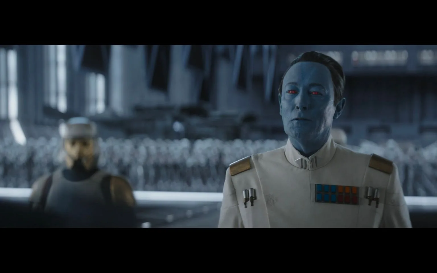 The image shows a scene from a "Star Wars" production featuring a character named Thrawn. He stands prominently in the foreground, characterized by his distinctive blue skin and red eyes. Thrawn is dressed in an Imperial officer's uniform, complete with rank insignia on the chest and epaulets on the shoulders. The background features a blurred figure of a helmeted Imperial soldier, suggesting a military setting within a large, structured hangar or assembly area filled with troops. The lighting is cool and somewhat dim, consistent with the regimented and austere aesthetic of the Empire in the "Star Wars" universe. The overall composition conveys a sense of authority and command, with Thrawn appearing calm and composed, reinforcing his role as a strategic and formidable leader within the narrative.