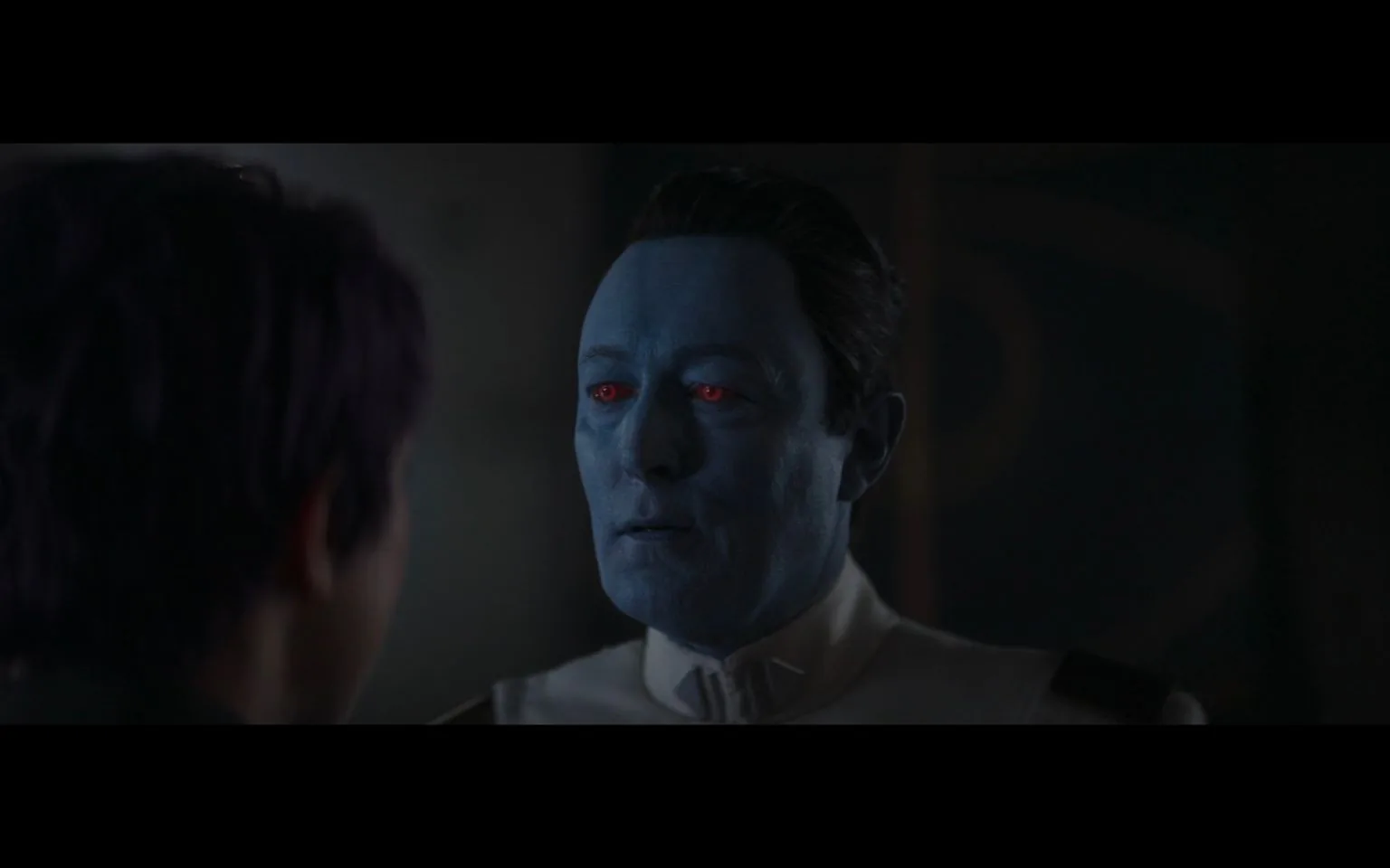 The image depicts a dramatic scene from the "Star Wars" universe. It features a character with blue skin and red eyes, dressed in a white uniform with black accents. The character appears to be engaged in a serious conversation with another person whose back is to the camera. The setting is dimly lit, adding to the tension of the encounter. The lighting highlights the blue-skinned character's facial features, emphasizing a calm yet intense expression. The composition is focused on the interaction between the two characters, with the blue-skinned character positioned slightly to the right, creating a sense of depth and focus on the dialogue between them. The scene has a somber and serious mood, typical of the dramatic storytelling in the "Star Wars" series.