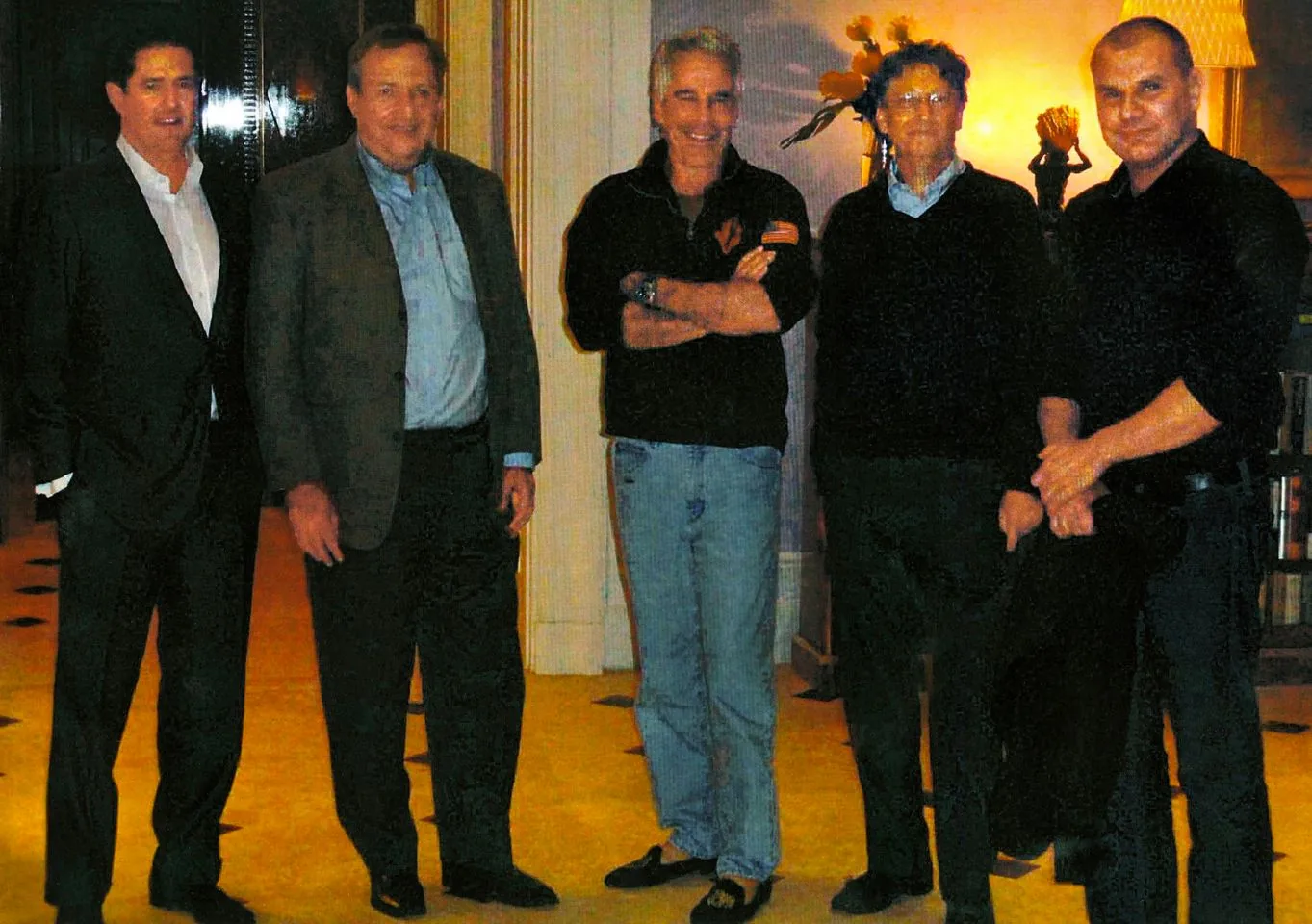 The image shows a group of five men standing closely together in an indoor setting. The individuals are dressed in a mix of business and casual attire, with some wearing suits and others in more relaxed clothing such as jeans and sweaters. The room has a warm and inviting atmosphere, with decorative elements like flowers and a lamp creating a homely ambiance. The lighting is soft, contributing to a relaxed mood. According to the hint provided, the individuals include Larry Summers, Jeffrey Epstein, and Bill Gates. The men appear to be engaged in a social gathering, possibly a private meeting or informal event. Each person displays a different posture, with some standing more formally and others adopting a casual, open stance. The background features subtle details like bookshelves and artwork, adding to the refined setting.