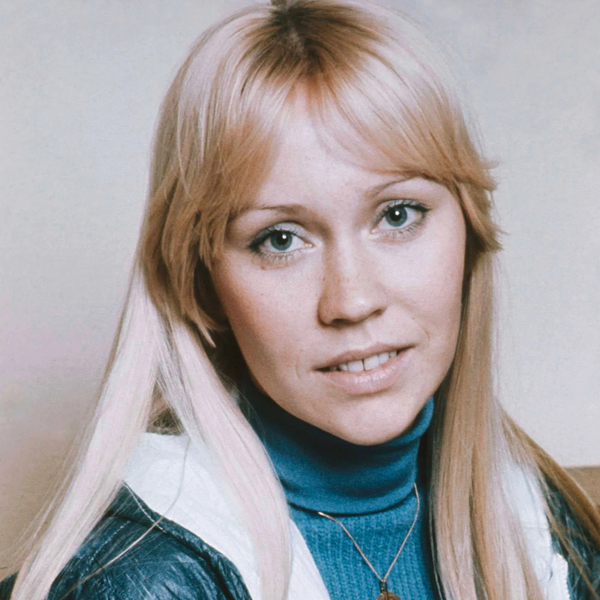 In this image, Agneta Åse "Agnetha" Fältskog is portrayed in a close-up shot, emphasizing her facial features. Her expression is calm and serene, with a slight smile that adds warmth to her demeanor. She has long, straight blonde hair that frames her face, and she wears a blue turtleneck sweater underneath a jacket, hinting at a casual yet stylish look typical of the 1970s. The lighting is soft and even, highlighting her clear complexion and light blue eyes. The background is neutral, focusing attention entirely on her face. This image appears to be taken indoors, likely in a studio setting, providing a clean and simple backdrop that complements her natural appearance.