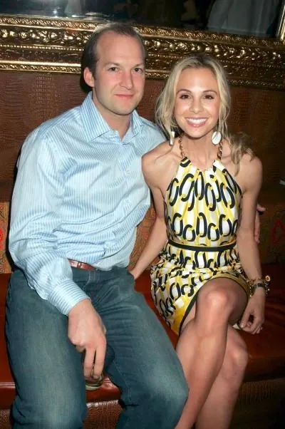 The image shows Tim and Elisabeth Hasselbeck sitting together in a cozy indoor setting. Tim is dressed in a light blue striped button-up shirt and jeans, holding a drink in his hand. Elisabeth is wearing a vibrant yellow dress with a bold black and white pattern, complemented by dangling earrings and bracelets, which add a touch of elegance to her attire. The couple is seated on a cushioned bench, with a decorative, ornate frame visible in the background, suggesting a warm and upscale ambiance. The lighting in the scene is soft, highlighting their relaxed and cheerful expressions as they pose closely together. The overall atmosphere is intimate and friendly, capturing a moment of togetherness.