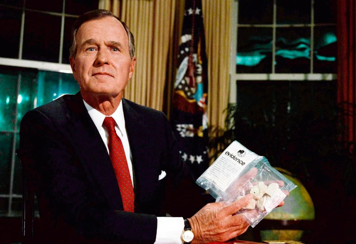The image shows HW Bush seated at a desk, holding a clear plastic evidence bag. The bag contains a substance and has the word "evidence" clearly printed on it. HW Bush is dressed in a formal dark suit with a white shirt and a red tie, exuding a serious and composed demeanor. The setting appears to be an official office, possibly the Oval Office, given the presence of heavy curtains and a globe in the background. A U.S. flag is partially visible, standing to the right. The lighting is warm, focusing on HW Bush, creating an authoritative and somber atmosphere. The image captures a moment likely related to a speech or announcement concerning drug policy or law enforcement, given the evidence bag's prominence.
