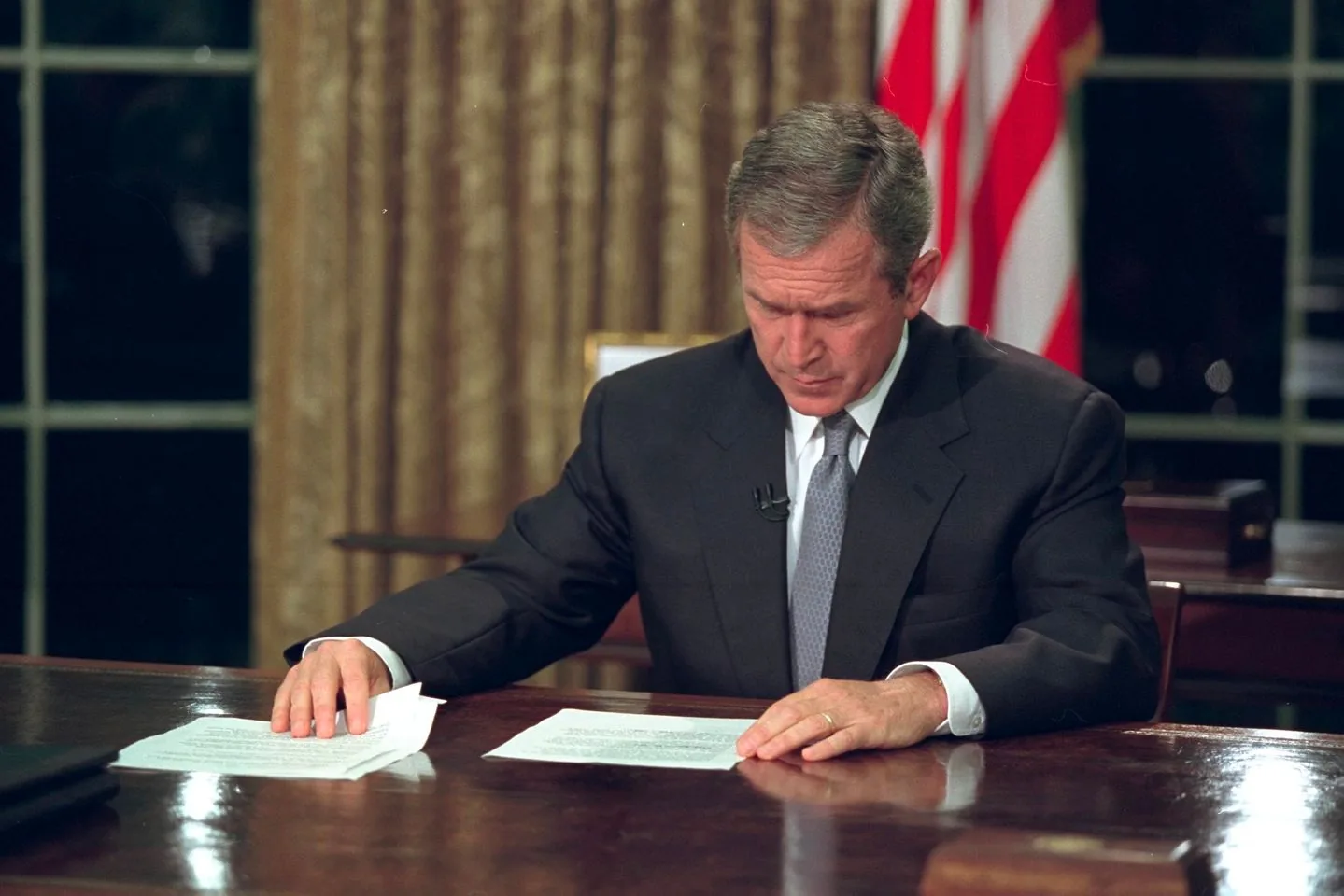 The image depicts President George W. Bush sitting at a desk, likely in an official setting such as the Oval Office. He is dressed in a dark suit with a white shirt and a light-colored tie, giving him a formal appearance. The background features heavy curtains and a part of an American flag, which suggests a setting of national importance. The lighting is soft but focused, illuminating the desk where President Bush is looking down at papers in front of him with a serious expression. This suggests he is reviewing important documents or preparing for an address. The desk is polished wood, adding to the formality of the scene. Behind him, the blurred outlines of furniture and office decor are visible, enhancing the atmosphere of an official workspace.