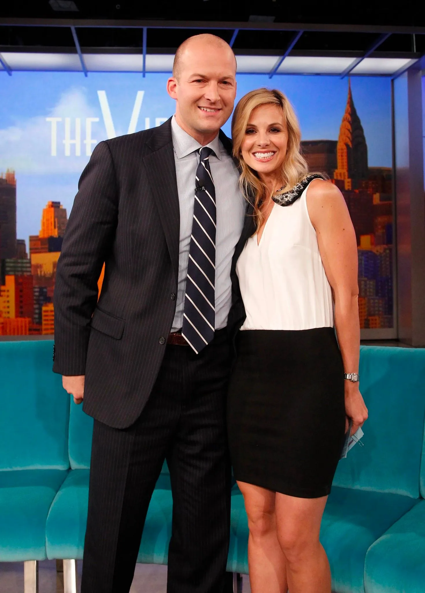 The image shows Tim and Elisabeth Hasselbeck on the set of a television show. They are standing close together in a studio setting, which is part of "The View," as indicated by the text in the background. Elisabeth Hasselbeck is wearing a stylish sleeveless dress with a white top and a black skirt, complemented by a decorative collar. Her hair is styled in loose waves, and she is smiling brightly at the camera. Tim Hasselbeck is dressed in a dark pinstripe suit with a white shirt and a striped tie, giving a formal appearance. The set features a turquoise couch and a backdrop depicting a city skyline, adding a vibrant and urban feel to the scene. The lighting is bright and even, typical of a professional studio environment, enhancing the cheerful and engaging atmosphere of the interaction.