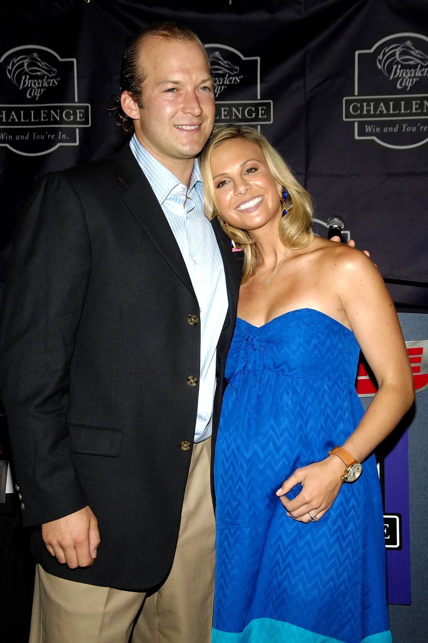 Tim and Elisabeth Hasselbeck are posing together at an indoor event. The backdrop features the "Breeders' Cup Challenge" logo, accompanied by the phrase "Win and You're In." Tim is dressed in a dark blazer over a light blue shirt and beige pants. He has a relaxed demeanor, with one arm around Elisabeth. Elisabeth is wearing a strapless blue dress with a subtle zigzag pattern and a watch on her wrist. Her expression is cheerful and affectionate as she leans into Tim. The lighting is bright, emphasizing their smiles and creating a warm, inviting atmosphere. This image captures a celebratory moment, likely at a formal or semi-formal occasion related to the Breeders' Cup event.