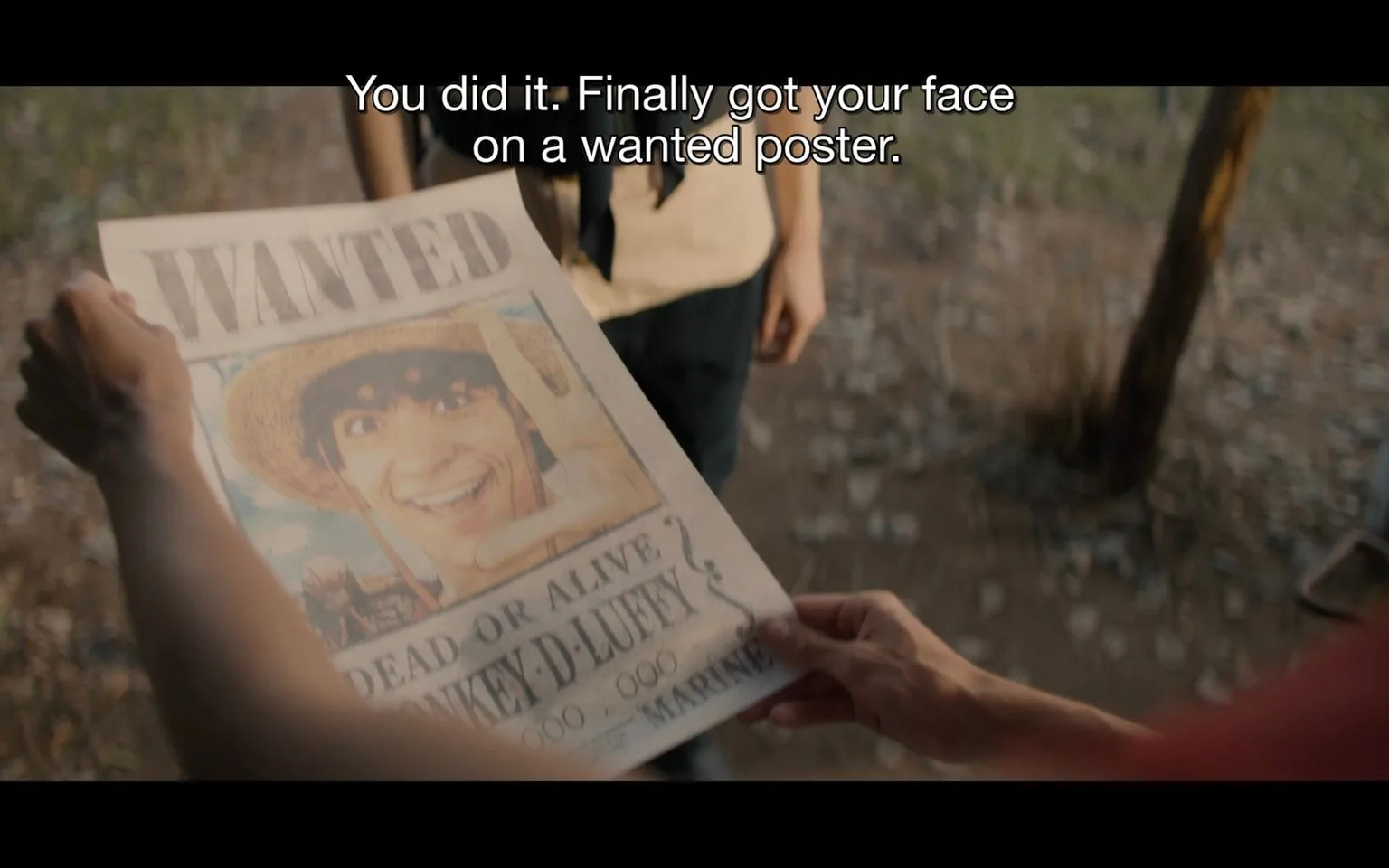 In this scene from "One Piece," an outdoor setting shows a person holding a wanted poster prominently featuring a character named Monkey D. Luffy. The poster, held up for another person to see, displays an image of Luffy with a big smile, wearing his iconic straw hat. The text on the poster includes "WANTED," "DEAD OR ALIVE," and "MONKEY D. LUFFY," indicating that Luffy is sought after by the authorities, as per the storyline of "One Piece." The background is blurred, suggesting a focus on the poster itself, with the ground covered in what appears to be dry earth or stones, hinting at a rural or deserted area. The lighting is natural, likely indicating daytime. The subtitles capture a moment of dialogue: "You did it. Finally got your face on a wanted poster," emphasizing a humorous or significant moment in the narrative. The overall mood is lighthearted, with a sense of adventure and camaraderie.