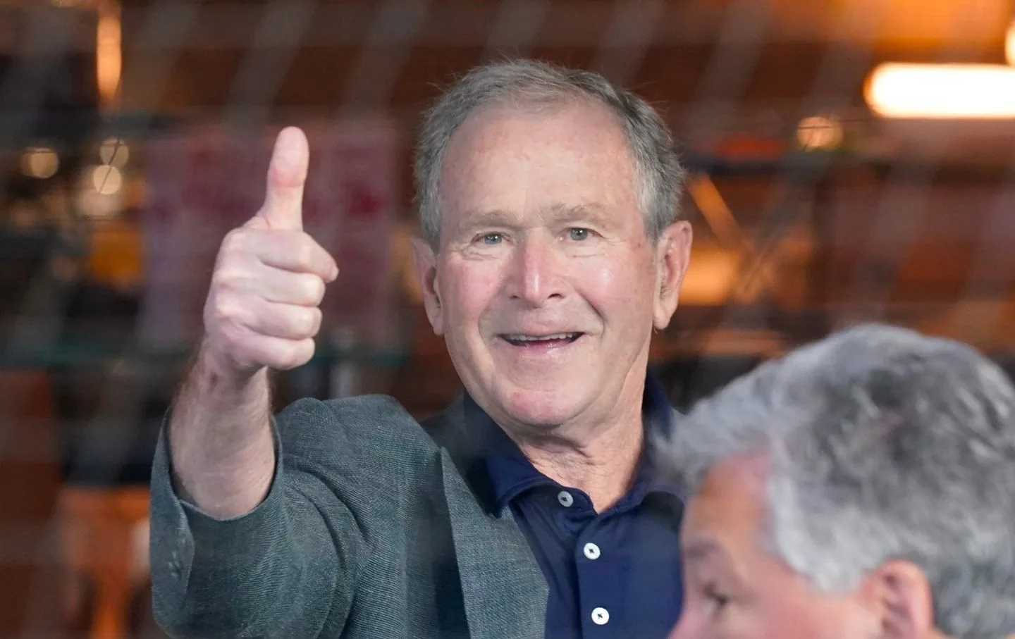 ```
President George W. Bush is depicted giving a thumbs-up gesture in this indoor setting. He is dressed in a dark blue polo shirt with a gray blazer. The expression on his face is friendly and cheerful, suggesting a positive or affirming mood. The background is slightly blurred, with warm lighting and some indistinct shapes, possibly indicating a casual or public event setting. Another person is partially visible in the foreground, but their identity is not discernible. The overall atmosphere of the image conveys an approachable and pleasant vibe, with the focus clearly on President George W. Bush's gesture and expression.