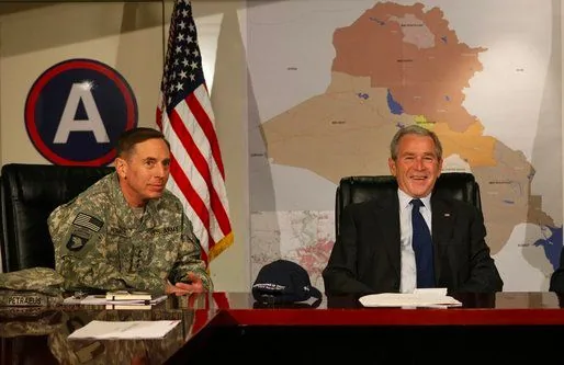 In the image, President George W. Bush and David Petraeus are seated at a table in what appears to be a formal setting. President Bush is dressed in a dark suit with a white shirt and tie, smiling broadly, creating a friendly and relaxed atmosphere. To his left, David Petraeus is wearing a military uniform, adorned with various badges and insignia, indicating his stature and experience in the military.
Behind them, an American flag is prominently displayed, alongside a large map of Iraq, which hints at the context of military and political discussions. The room has a formal and official ambiance, with a logo featuring a large "A" visible on the wall, possibly indicating a military command or unit.
The table in front of them is neat, with a cap and some documents placed on it. The lighting is bright, emphasizing the importance of the meeting and ensuring clarity in the photograph. The image captures a moment of camaraderie and professionalism, underscoring the significance of their roles in the context of international affairs.