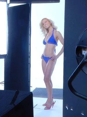 Miss Elisabeth Hasselbeck is featured in a studio photoshoot. She is wearing a vibrant blue bikini, which contrasts with her blonde hair, adding a vivid pop of color to the scene. The setting appears to be a professional photography studio with lighting equipment visible around her, which contributes to a bright, well-lit environment. Miss Hasselbeck stands confidently with her hands on her hips, showcasing a poised and assured posture. Her expression is pleasant, suggesting a relaxed and professional demeanor. She is wearing high-heeled sandals that further complement her attire. The background is predominantly white, likely to enhance the focus on her and create a clean aesthetic. The overall composition is polished, indicating a carefully arranged photoshoot setup.