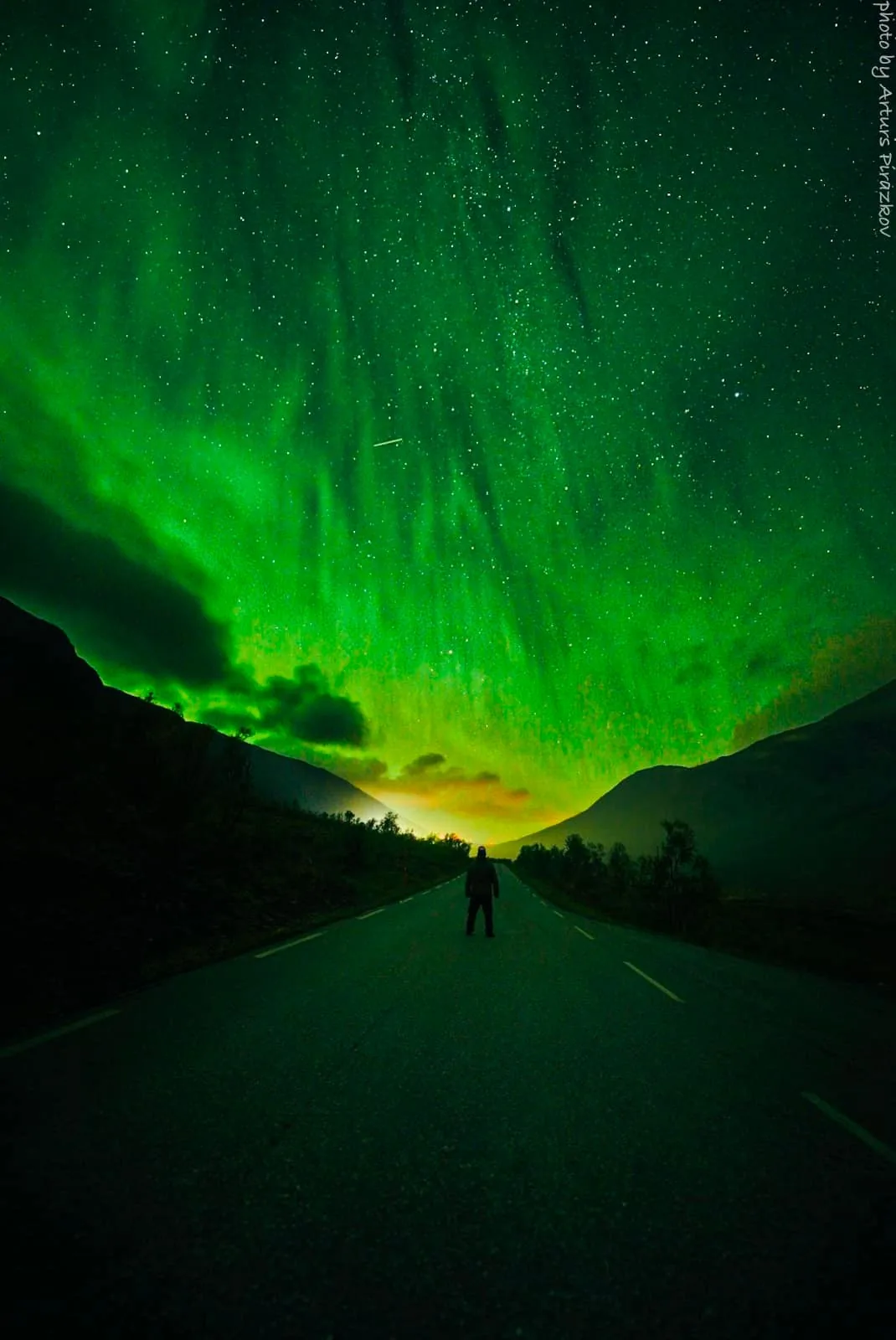 The image features a captivating view of the aurora borealis, commonly known as the Northern Lights, swirling across the night sky in vibrant shades of green. A person stands in the middle of a deserted road, silhouetted against the mesmerizing backdrop. This individual appears to be observing the natural spectacle, emphasizing a sense of solitude and wonder. The road leads into the distance, flanked by dark, shadowy hills that frame the scene. The sky is filled with countless stars, enhancing the magical atmosphere. This photograph by Arturs Pážokov captures the raw beauty of nature, with the aurora’s luminescence providing the primary source of light. The overall mood is serene and awe-inspiring, showcasing the grandeur of the natural world.