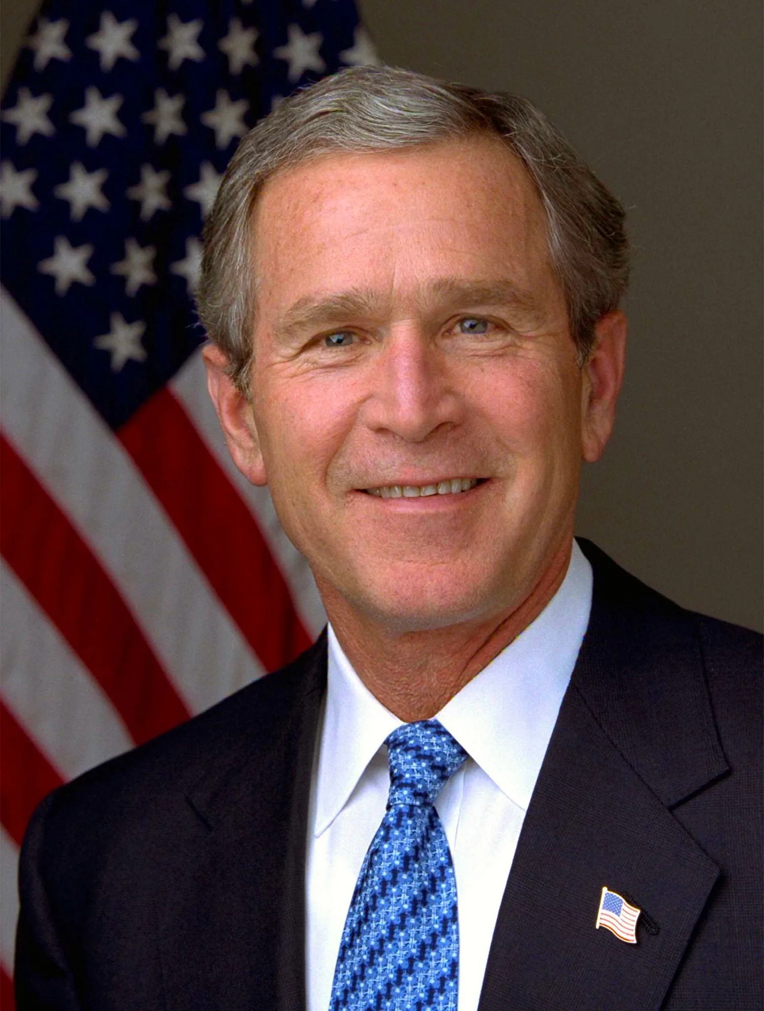 The image is a formal portrait of President George W. Bush. He is positioned in front of a blurred American flag, suggesting a patriotic and official setting. The composition is typical of a presidential portrait, with President Bush centered in the frame, conveying a sense of authority and presence. He is wearing a dark suit jacket paired with a white dress shirt and a blue patterned tie. A small American flag pin is visible on the lapel of his jacket, reinforcing the patriotic theme. The lighting is even and bright, highlighting his facial features and giving the portrait a polished and professional appearance. There is a calm and composed expression on his face, typical of official portraits, aiming to convey confidence and leadership. This image likely served as an official representation during his time in office.