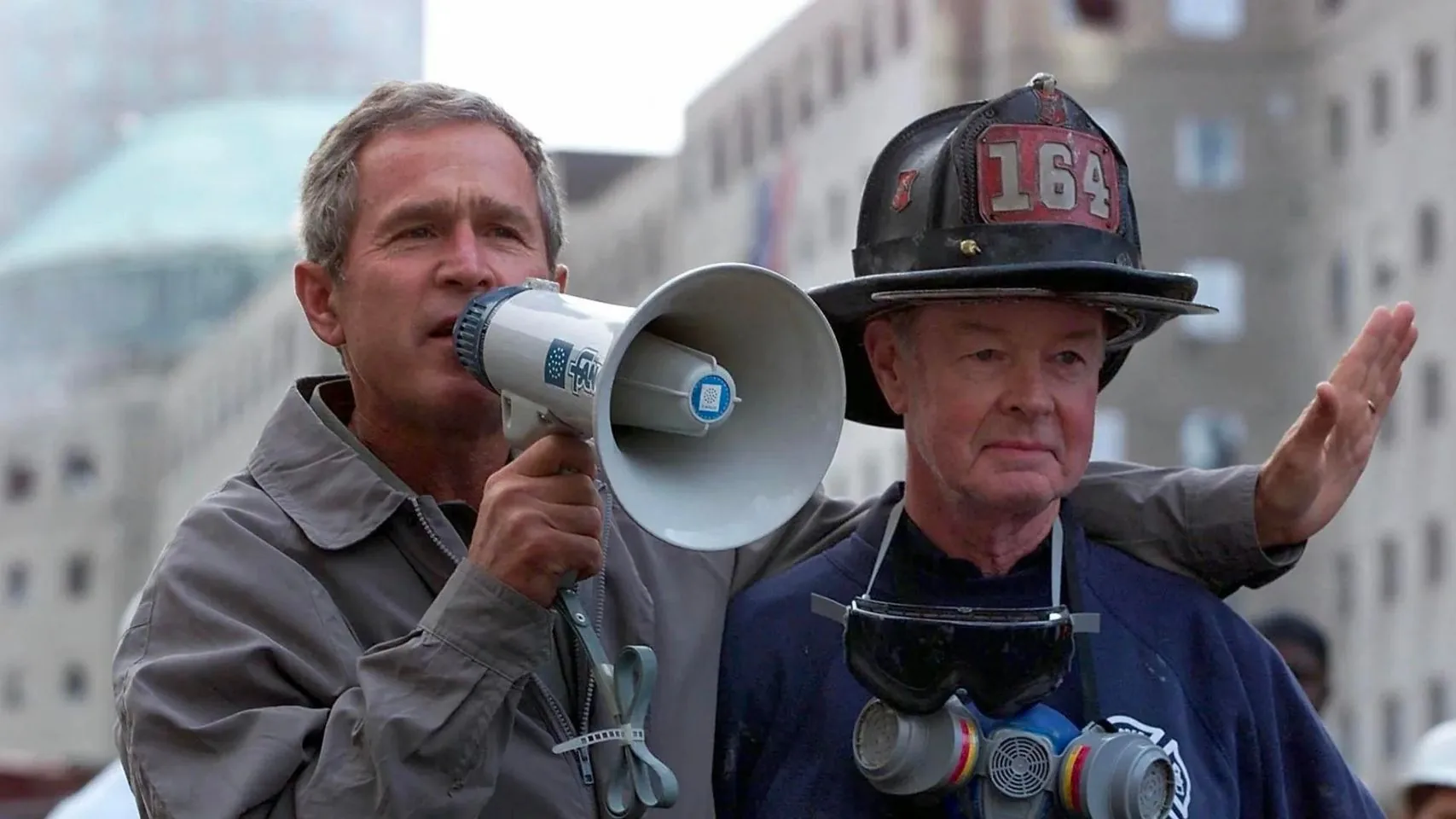 President George W. Bush is pictured in this historical photo from 9/11, standing amidst the rubble in a city setting. He holds a megaphone in his hand, symbolizing a moment of direct communication and leadership. Beside him stands a firefighter wearing a helmet marked with the number 164 and a respirator hanging from his neck, reflecting the emergency response efforts. The backdrop consists of tall buildings, giving a sense of the urban environment that was central to the tragic events of 9/11. President Bush has his arm around the firefighter, demonstrating solidarity and support during this critical moment. The lighting is natural, emphasizing the urgency and gravity of the scene. The attire of both individuals is practical and suited for the conditions, with Bush wearing a jacket suitable for outdoor activities. This image captures a poignant instance of leadership and unity in the face of disaster.