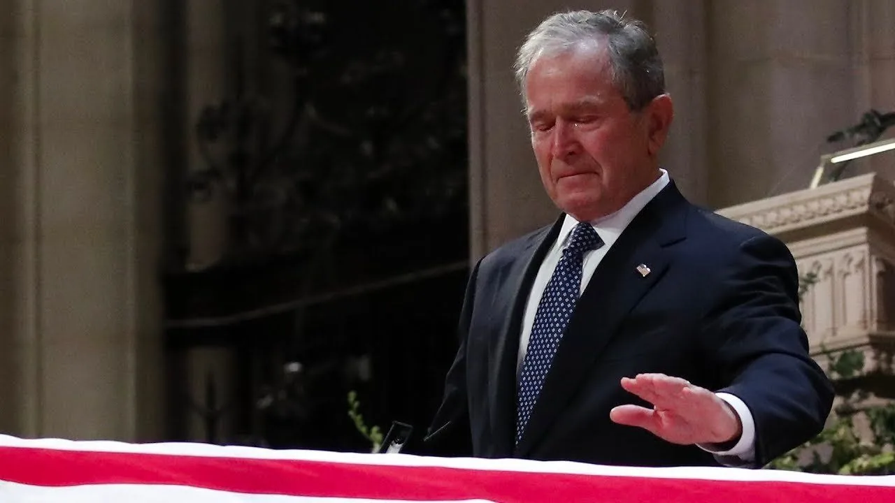President George W. Bush is depicted in this image standing solemnly at a ceremony. He is in an indoor setting, likely a church or cathedral, given the ornate architecture visible in the background. The lighting is subdued, befitting a somber occasion. President Bush is wearing a dark suit with a white shirt and a patterned tie, appropriate attire for a formal event. He appears to be in a moment of reflection or mourning, as his expression conveys sadness and contemplation. In the foreground, there is a flag-draped casket, indicating this is part of a funeral service. The flag is prominently displayed, and its patriotic colors stand out against the more muted tones of the surroundings. The image captures a poignant moment, emphasizing the personal and national significance of the event.