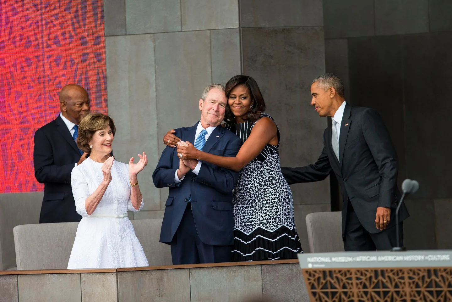 The image captures a significant moment at the opening of the National Museum of African American History and Culture. In the foreground, Michelle Obama is warmly embracing President George W. Bush. They both appear joyful, symbolizing unity and camaraderie. Michelle Obama wears a black and white patterned dress, while President George W. Bush is dressed in a dark suit and blue tie.
To their left, Laura Bush claps, wearing a white dress, expressing appreciation and support. In the background, another figure in a suit is smiling, contributing to the celebratory atmosphere. President Barack Obama is seen extending his hand, enhancing the sense of connection and significance of the event.
The backdrop features a textured wall, adding depth to the composition. The lighting is bright and clear, typical of an outdoor event. The text "NATIONAL MUSEUM AFRICAN AMERICAN HISTORY CULTURE" suggests the location and occasion, marking this as a notable historical event.