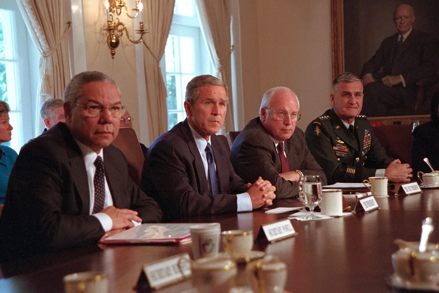 In this image, several high-profile political figures are seated at a large wooden conference table during a formal meeting. The setting is indoors, likely a government office or a similar official venue, characterized by elegant decor, including a chandelier and draped curtains, suggesting a formal and authoritative atmosphere.
Colin Powell, President George W. Bush, and Dick Cheney are prominently seated in the foreground. Colin Powell, wearing a dark suit and polka dot tie, appears attentive and serious. President George W. Bush, also in a dark suit with a blue tie, looks focused and engaged, conveying the gravity of the discussion. Dick Cheney, sitting next to Bush, wears glasses and a dark suit, exuding a composed demeanor.
Another military official in uniform is seated beside Cheney, adding a sense of authority and formality to the scene. The table is set with cups, saucers, and nameplates, emphasizing the structured and professional nature of the meeting. The lighting is warm, highlighting the participants and adding to the serious yet respectful mood of the gathering. A portrait hangs in the background, contributing to the historical and formal context of the setting.