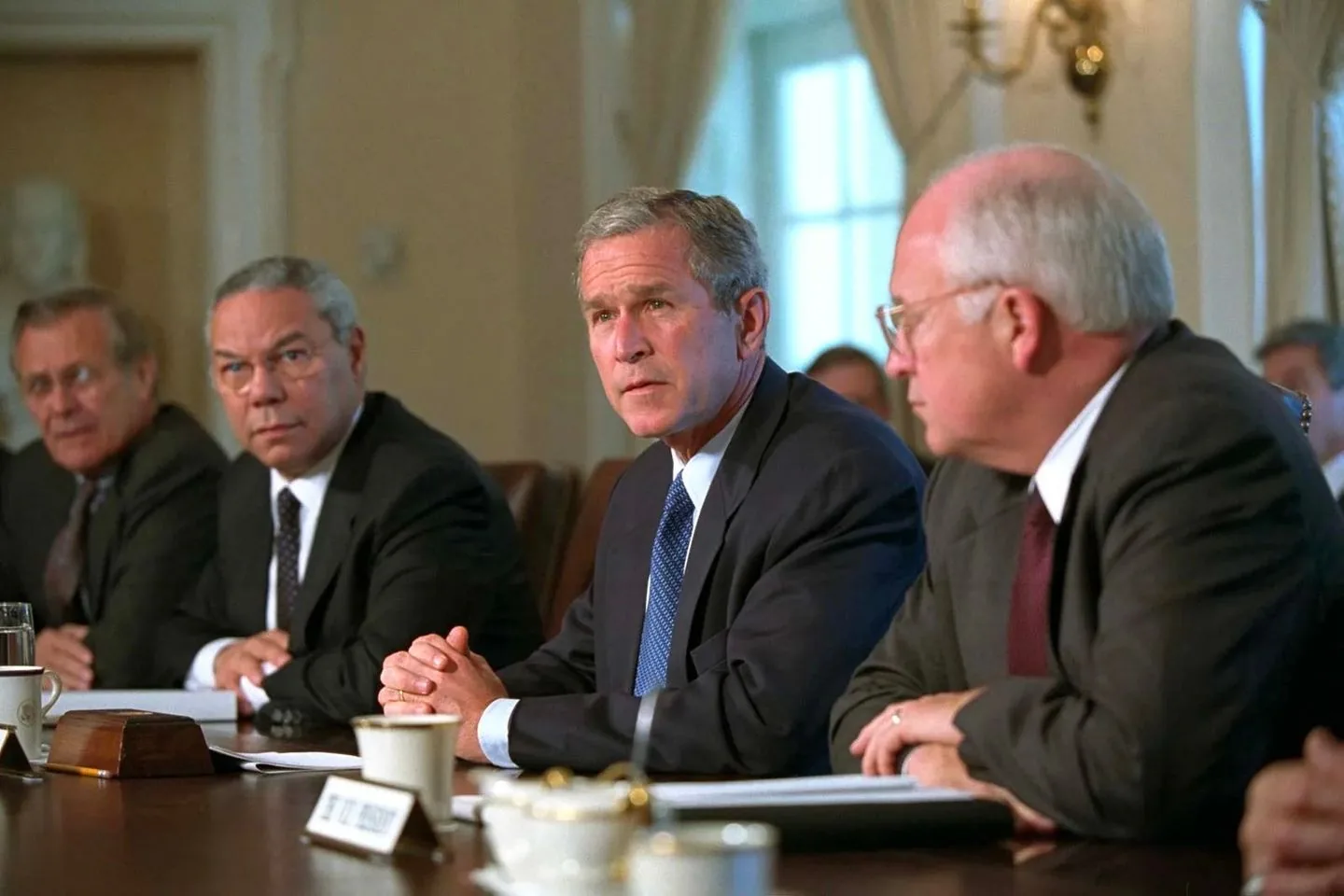 The image depicts a formal meeting setting, likely in a government or political environment. Seated at a long table, from left to right, are Colin Powell, President George W. Bush, and Dick Cheney. The room has a dignified atmosphere with cream-colored walls and traditional decor, including a bust visible in the background. Natural light filters through large windows, casting a soft glow over the scene.
Colin Powell, wearing a black suit and polka-dot tie, looks attentive and engaged. President George W. Bush is centered in the image, dressed in a dark suit and blue tie, with his hands clasped on the table, exuding a focused demeanor. Dick Cheney is on the right, also in a dark suit, with his profile visible as he listens intently.
The table is adorned with white coffee cups and notepads, suggesting a formal discussion or briefing. Nameplates are visible but blurred, adding to the official nature of the gathering. The mood is serious, indicating the discussion of significant matters. The lighting is soft and the overall composition is well-balanced, highlighting the importance of the individuals and the meeting.