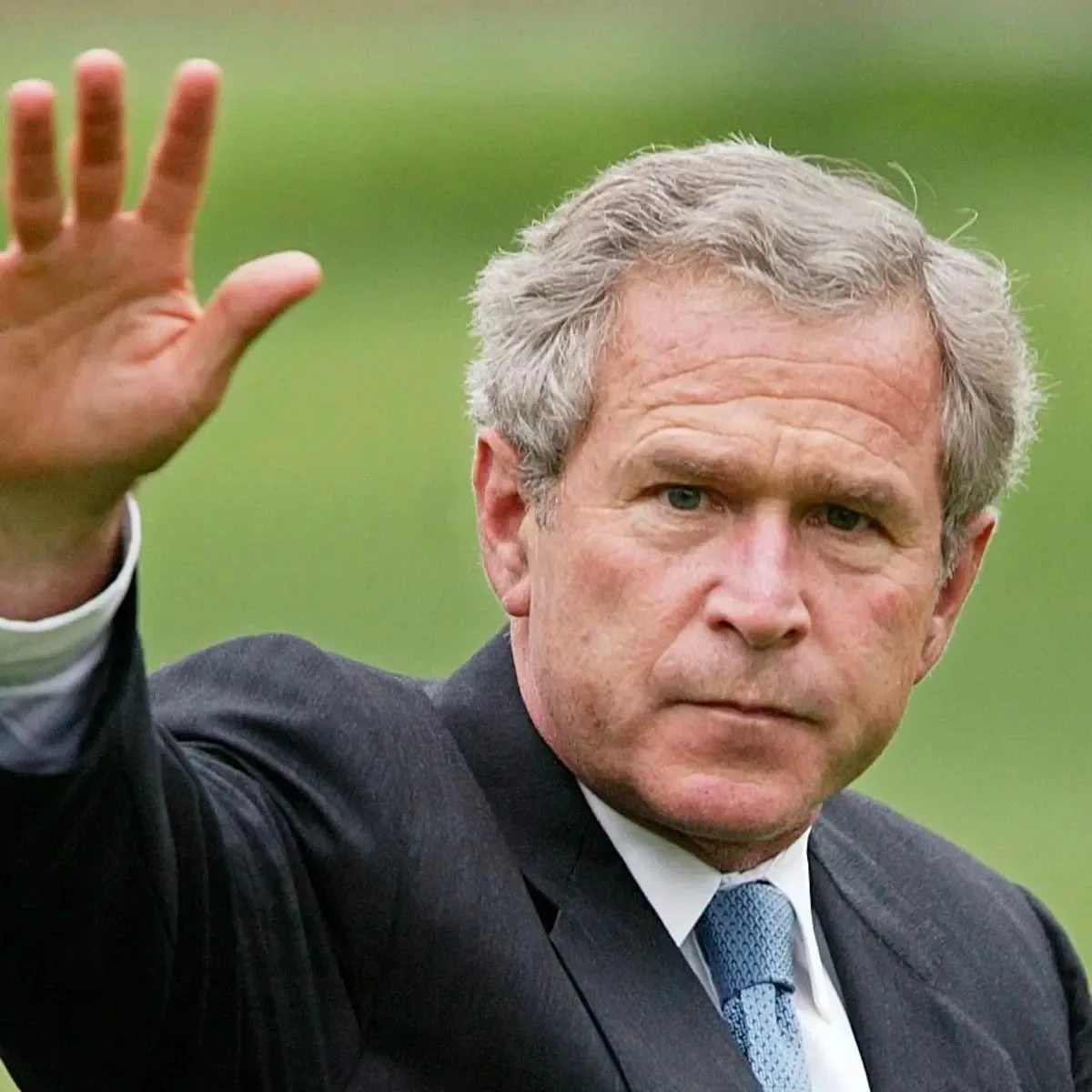 The image features President George W. Bush, captured in an outdoor setting. He is in the foreground, wearing a dark suit with a light blue tie, typical of formal political attire. His expression is serious, emphasizing a moment of solemnity or contemplation. He is raising his right hand, possibly in a wave or a gesture of acknowledgment. The background is a blur of greenery, suggesting he might be in a park or a garden setting, which adds a serene contrast to his formal demeanor. The lighting is natural, indicating that the photo was taken during the daytime. This image captures a moment of political life, reflecting the responsibilities and public interactions of a president.
