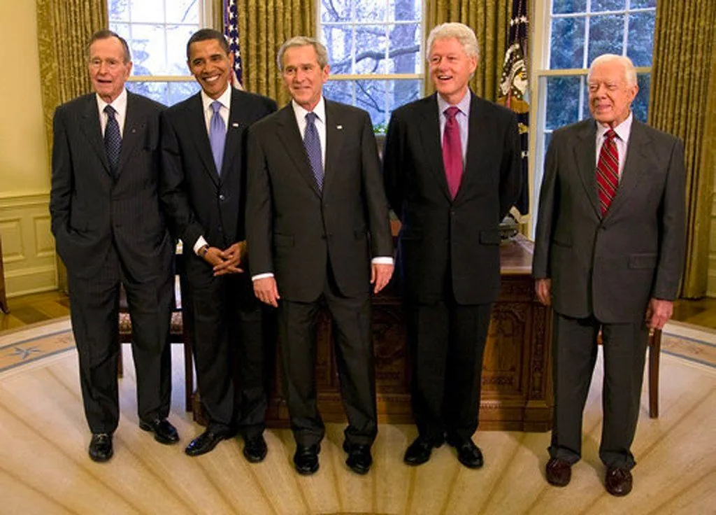 The image depicts a formal gathering in what appears to be the Oval Office, indicated by the presidential setting and the presence of the Resolute Desk in the background. The scene includes five men dressed in business attire, standing in a line and posing for a photograph. From left to right, the individuals are identified using the provided hint as hw, bho, gw, bill clinton, and jimmy clinton. They are all wearing dark suits, with white shirts and variously colored ties, adding a sense of formality and cohesion to the group. The mood is professional yet friendly, as all the individuals are smiling. The lighting is bright, suggesting the photograph was taken during the day. Behind them, large windows with drawn curtains let in natural light, and the American flag is visible, enhancing the official and patriotic atmosphere. The carpet features a subtle design, possibly with symbolic motifs. Overall, this image captures a moment of camaraderie and historical significance among prominent figures.