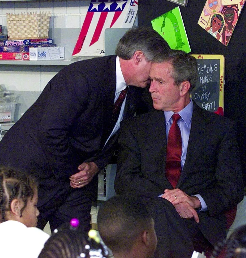 President George W. Bush is seated in a classroom setting, dressed in a dark suit with a blue shirt and red tie. His posture is attentive and serious as he listens to a man whispering in his ear. The man is wearing a dark suit and leaning in close to President Bush, suggesting an urgent or important exchange. The background features educational materials, including a blackboard with the text "READING MAKES A COUNTRY GREAT!" and various children's drawings. The mood in the room appears tense, likely due to the serious nature of the information being conveyed. The classroom setting is further emphasized by the presence of young students seated in the foreground, their faces partially visible. The lighting is bright and typical of an indoor educational environment, highlighting the expressions and interactions of the individuals present.