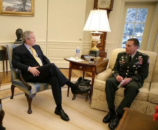 In the image, President George W. Bush is seated in a formal office setting, possibly the Oval Office, engaging in conversation with General Petraeus. President Bush is sitting on a striped upholstered chair, dressed in a dark suit with a light blue shirt and a yellow tie, exuding a formal and professional demeanor. His posture is relaxed, with one leg crossed over the other, and his hands resting comfortably in his lap as he listens attentively.
To his right, General Petraeus is seated in a similar chair, dressed in a military uniform adorned with various medals and insignia, reflecting his high rank and accomplishments. He holds what appears to be a document or folder in his hands, suggesting a discussion or briefing. The room is warmly lit, with a tall lamp casting light over a wooden table positioned between them, which holds a phone and a glass of water. Behind them, a window reveals a glimpse of the outside environment, indicating it might be daytime. The setting is formal and suggests a high-level meeting or briefing.