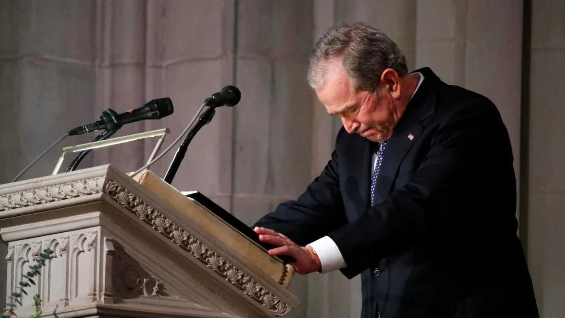 President George W. Bush is depicted in a solemn moment at a pulpit, suggesting he is likely delivering a speech or reflecting during a significant event. He is dressed in a formal dark suit, white shirt, and patterned tie, embodying a dignified appearance suitable for a formal occasion. The setting is an indoor environment, possibly a church or a formal hall, indicated by the ornately carved pulpit and the presence of microphones mounted at the top. The lighting is soft, creating a respectful and contemplative atmosphere. President Bush's posture is slightly bent forward, with his hands resting on the pulpit, conveying a sense of introspection or solemnity. This image captures a poignant moment, emphasizing sincerity and gravity. The background is neutral, directing attention to his expression and the context of the event.