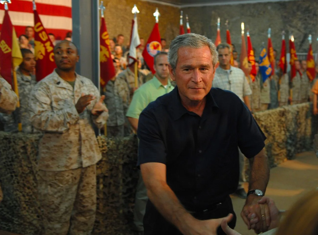 The image captures President George W. Bush at what appears to be a military event. He is in the foreground, reaching out to shake hands with someone out of frame, displaying a serious yet approachable demeanor. He wears a dark short-sleeved shirt, contrasting with the lighter camouflage uniforms of the military personnel around him. In the background, several soldiers stand in formation, some clapping, while others hold flags that are prominently displayed. The flags are a mix of red and yellow with various insignias, adding a vibrant backdrop to the scene. The setting is indoors, with a camouflaged netting draped over the walls, contributing to the military ambiance. The lighting is warm, casting a golden hue over the scene, which enhances the formal yet intimate atmosphere of the event. The soldiers' attentive postures indicate that this could be an official ceremony or speech. The focus on President George W. Bush emphasizes his central role in the event.