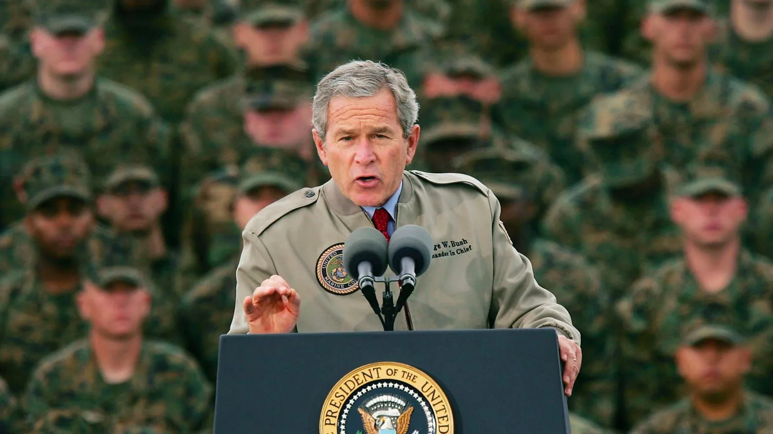 1) Rich description: The image depicts President George W. Bush delivering a speech outdoors, surrounded by military personnel in uniform. He stands at a podium labeled with the "President of the United States" seal, suggesting a formal and official event. President Bush is wearing a light-colored jacket with an emblem and his name, "George W. Bush," along with the title "Commander in Chief" embroidered on it. His expression is focused and earnest as he addresses the audience. The background is filled with uniformed military personnel who are attentively observing the speech, creating a solemn and respectful atmosphere. The lighting is natural, indicating the event takes place during the day. The composition emphasizes the President at the center, highlighting his role as a speaker and leader.
2) On-screen text block:
- Title: "..."