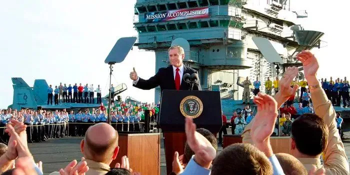 President George W. Bush is seen delivering a speech on the deck of a naval ship, with a prominent "MISSION ACCOMPLISHED" banner displayed in the background. He is standing at a podium bearing the presidential seal, gesturing with a thumbs-up, which adds an optimistic tone to the setting. The crowd in the foreground, likely including military personnel, is applauding enthusiastically. The backdrop of the image features a large aircraft carrier, emphasizing the military context of the event. Various crew members in colorful uniforms are lined up on the ship, adding a sense of formality and organization. The lighting is bright, indicating a day-time outdoor setting, and the overall atmosphere is celebratory. President Bush is dressed in a suit, reinforcing the formal nature of the occasion. The scene captures a significant moment with a blend of political leadership and military achievement.