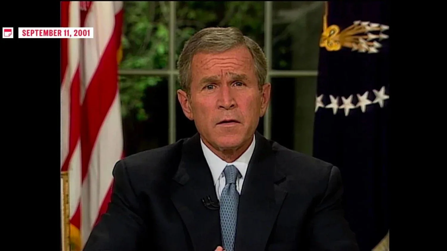 President George W. Bush is depicted in the image, delivering a speech. The setting is formal, likely from the Oval Office, indicated by the American flag and presidential seal partially visible in the background. Bush is dressed in a dark suit, white shirt, and a light blue tie, exuding a formal and somber demeanor. His expression is serious and focused, reflecting the gravity of the situation being addressed. The lighting is even and professional, typical of a televised address. The image prominently features the date "September 11, 2001," which is the context of his speech, addressing the nation following the tragic events of that day. The environment is official and composed, designed to convey a message of leadership during a crisis. The visual composition centers President Bush behind a desk, with his hands clasped, adding to the solemn atmosphere.
