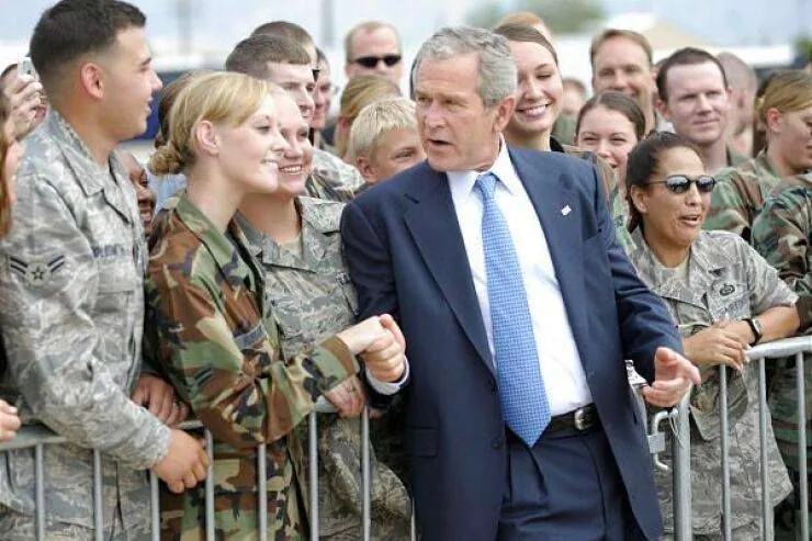 President George W. Bush is seen engaging with a group of military personnel. The setting is outdoors, likely on a military base or similar environment. President Bush is wearing a dark suit with a light blue tie, and he appears to be shaking hands and interacting warmly with the service members who are dressed in various military uniforms, including camouflage and digital patterns.
The mood is lively and positive, with several people smiling and appearing eager to meet the President. A barrier separates President Bush from the crowd, suggesting this is a formal visit or event. The lighting is natural, indicating it is daytime, and the background shows more people gathered, likely to see the President. The scene captures a moment of connection and mutual respect, typical of a presidential visit to military personnel.