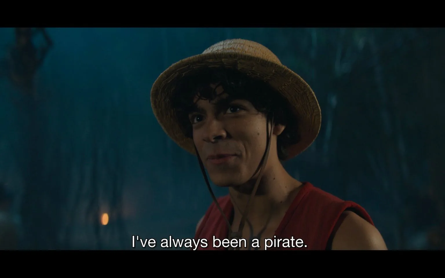 In this scene from the live-action adaptation of "One Piece," a character named Luffy is shown wearing a straw hat and a sleeveless red vest, which are iconic to his character. The setting appears to be outdoors at night, with a dimly lit background that suggests a mysterious or adventurous atmosphere. Luffy's expression is confident and determined as he declares, "I've always been a pirate." The lighting highlights his face, drawing attention to his earnest demeanor. This moment captures the essence of Luffy's adventurous spirit and unwavering commitment to his pirate identity. The colors are mostly muted, focusing on earthy tones that blend with the night scene, enhancing the adventurous theme of the series.