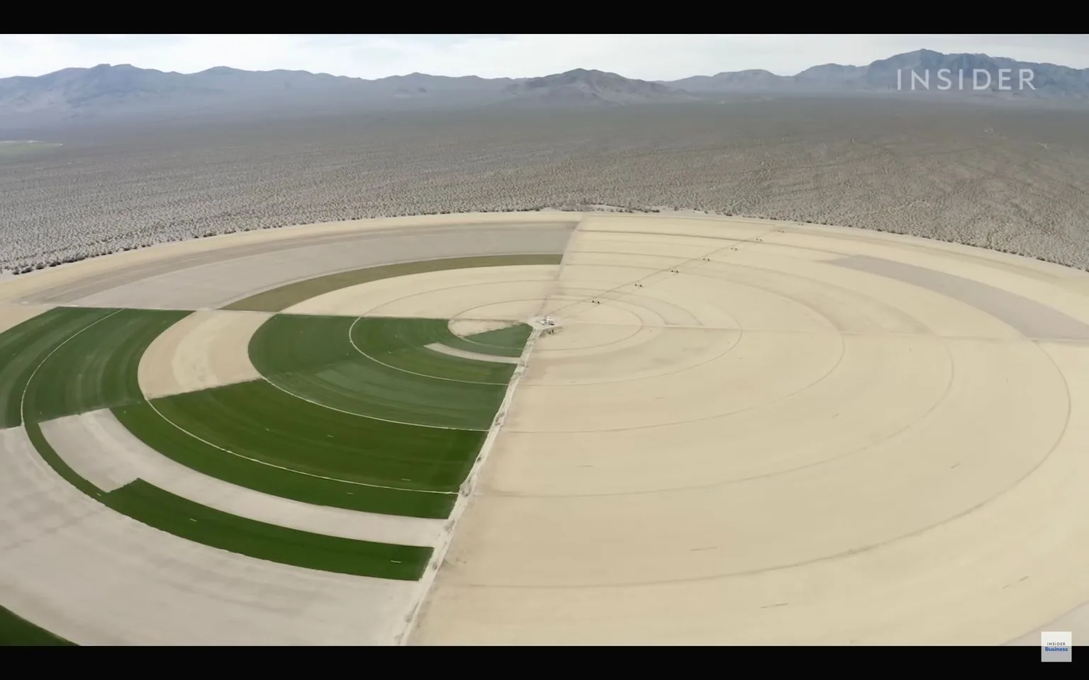 The image depicts a large circular agricultural field, captured from an aerial perspective. This field is located in a desert environment, with arid, mountainous terrain visible in the background. The field is divided into segments, with some areas showing vibrant green crops, likely irrigated, while other sections remain dry and barren. This pattern suggests the use of center pivot irrigation, a technique commonly employed to efficiently water crops in dry regions.
The contrast between the green and beige sections is visually striking, emphasizing the division between cultivated and uncultivated land. The landscape is vast, stretching towards the horizon, accentuating the isolation of the agricultural plot within the expanse of desert.
In the top right corner, the text "INSIDER" is visible, and at the bottom right, "Insider Business" is noted, indicating the source of the image. The lighting is bright and natural, typical of a sunny day, enhancing the colors and details in the scene. This setting reflects themes of innovation in agriculture and resource management in challenging environments.