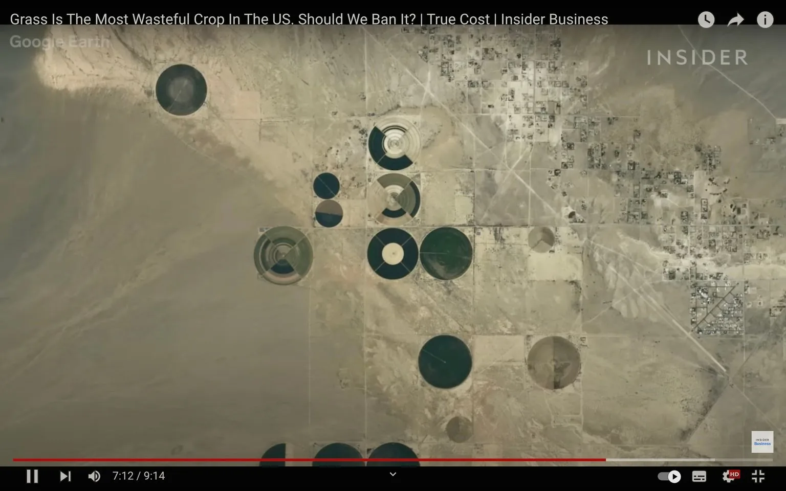 The image is a video frame from a YouTube video titled "Grass Is The Most Wasteful Crop In The US. Should We Ban It? | True Cost | Insider Business" on the Insider channel. It shows an aerial view of a desert landscape featuring numerous circular fields, likely created by center pivot irrigation systems. The area appears arid, with sandy and barren land surrounding the circles of vegetation.
In the upper left corner, "Google Earth" is visible, indicating the use of satellite imagery. The circular patterns vary in size and color, indicating different stages of cultivation or types of crops. Some circles are lush and green, while others appear more barren or have concentric patterns suggesting different growth phases or irrigation levels.
The scene highlights the contrast between the cultivated land and the surrounding desert, emphasizing the agricultural practice in a challenging environment. The setting's arid nature and the use of irrigation suggest a focus on water usage and sustainability, aligning with the video's theme of examining the environmental cost of cultivating grass in the U.S.
