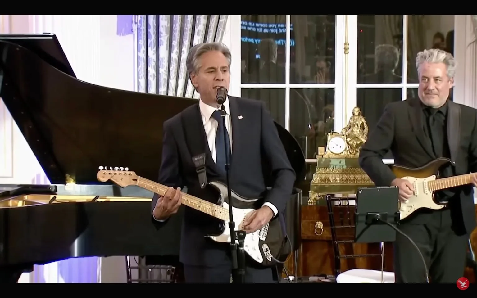 In this image, Tony Blinken is performing indoors at an event. He is playing an electric guitar and singing into a microphone. Dressed in a formal dark suit with a white shirt and a dark tie, he appears focused on his performance. Beside him is another musician also playing an electric guitar, dressed in a black suit with a black shirt and tie. The setting includes a grand piano to the side and a decorative antique clock on a wooden cabinet in the background, adding an elegant touch to the scene. The lighting is warm and the atmosphere suggests a formal gathering or diplomatic event. The image is a screenshot from a video published by The Independent on YouTube.