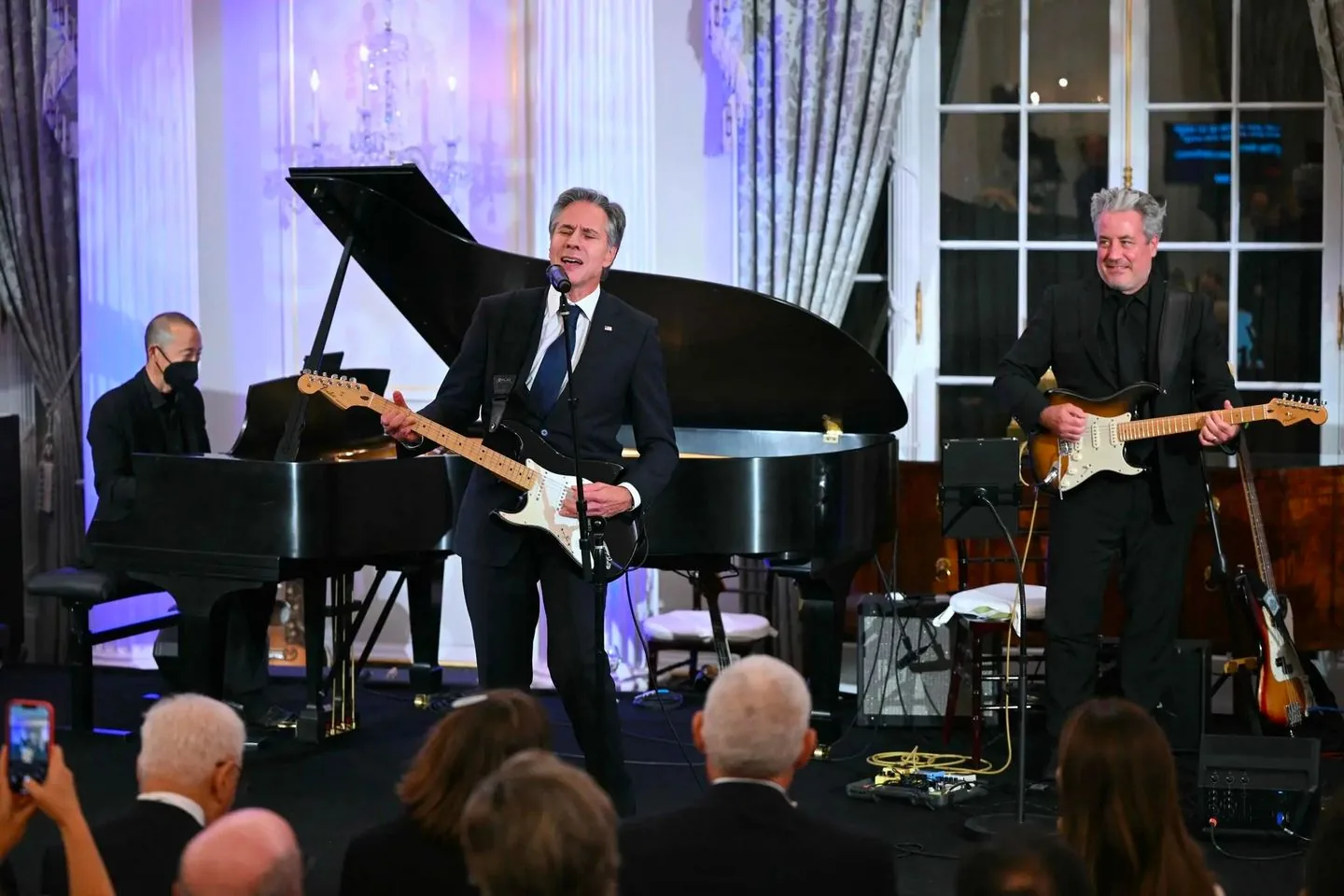 A live music performance is taking place indoors featuring Tony Blinken. The setting is elegant, with a grand piano and a crystal chandelier adding to the sophisticated ambiance. Tony Blinken is in the center, passionately playing an electric guitar and singing into a microphone. He is dressed in a formal dark suit, which contrasts with the vibrant energy of the performance. To his left, a pianist wearing a mask plays a grand piano, adding depth to the music. On the right, a guitarist accompanies Tony Blinken, also dressed in a black suit. The audience, seen from the back, is seated and appears engaged, with some capturing the moment on their phones. The room is well-lit, emphasizing the performers and creating a warm atmosphere, with curtains and windows adding to the refined setting.