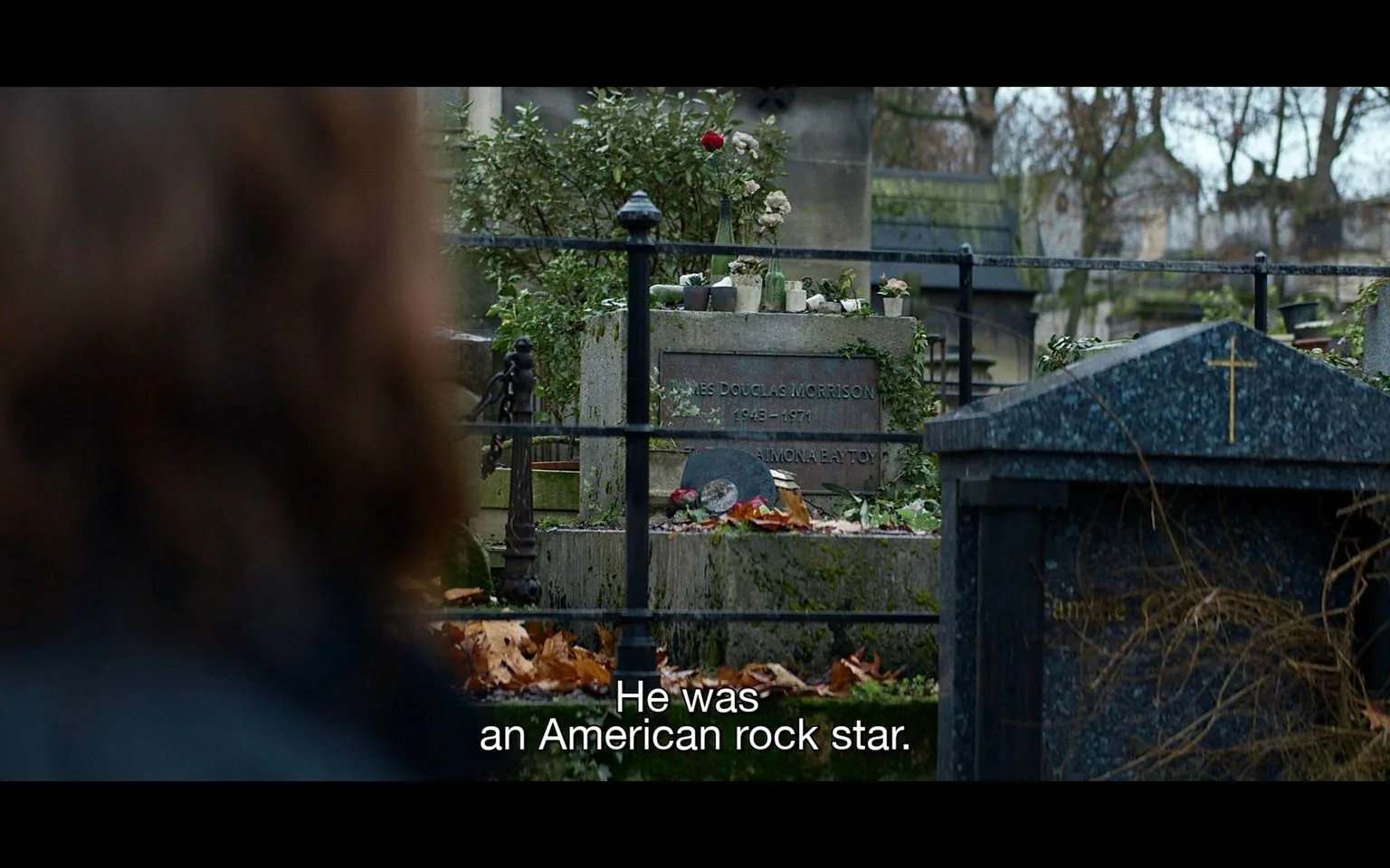 This image shows the grave of Jim Morrison in France. The setting is an outdoor cemetery, with the grave surrounded by metal railings and foliage suggestive of autumn, as leaves cover the ground. The gravestone has inscriptions reading "JAMES DOUGLAS MORRISON" and "1943 - 1971," indicating his years of birth and death, along with another line that is partially obscured. The grave is adorned with various offerings, including small potted plants, bottles, and a vinyl record, indicating Morrison's influence as a musician. In the foreground, the blurred silhouette of a person is partially visible, watching the grave. The lighting is natural and subdued, fitting the somber, reflective mood of the scene. The subtitles at the bottom of the image read, "He was an American rock star," reinforcing Jim Morrison's identity and legacy as a prominent figure in rock music.