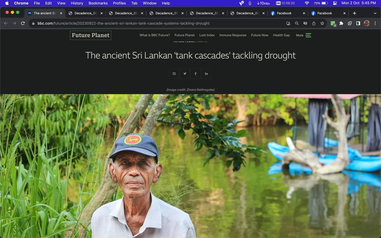 The image is a screenshot from an article on BBC's website, featuring the headline "The ancient Sri Lankan 'tank cascades' tackling drought." The background is a lush, natural environment with greenery and a body of water, suggesting a serene and verdant setting. In the foreground is an older man wearing a cap with a distinctive red and yellow emblem. His expression is calm and contemplative, possibly indicating his connection or relevance to the article's subject. The lighting is natural, highlighting the vibrant greens and blues of the surroundings. In the background, a blue boat with a canopy is visible, adding to the sense of tranquility and utility of the area. The image is credited to Zinara Rathnayake. The site or app is clearly identified as bbc.com, with the publisher being BBC. The article seems to focus on traditional methods of water management in Sri Lanka, which are being used to address modern drought challenges.
