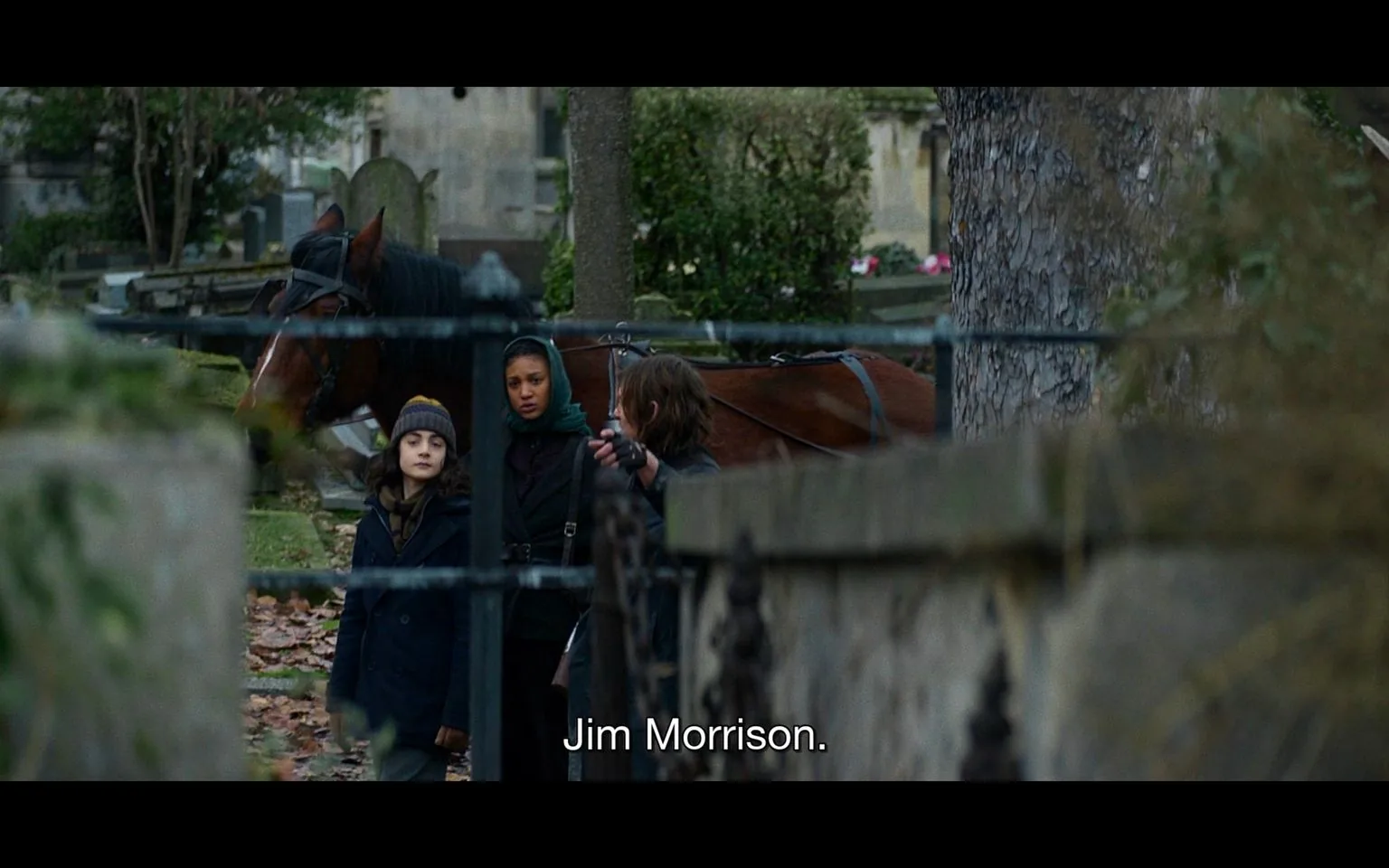 The scene depicts three individuals standing in a cemetery. They are near a horse, which is saddled, indicating they may have traveled on horseback. The setting features gravestones and trees, typical of a serene, historical cemetery. The individuals are dressed in winter clothing, suggesting cold weather. One person wears a beanie, another a scarf, and the third has gloves, emphasizing the seasonal chill. The atmosphere is somber and reflective, fitting for the setting. The background is blurred, focusing the viewer on the characters and their interaction. The caption "Jim Morrison." is displayed at the bottom, hinting at a discussion about visiting a notable grave. The colors are muted, adding to the sober tone.