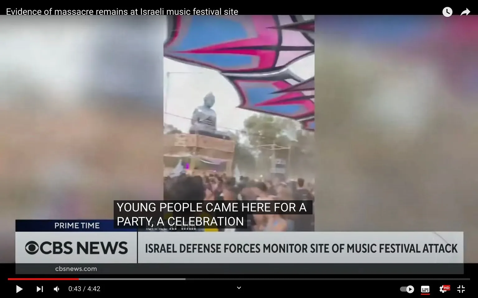 The video frame shows a scene from a music festival in Israel. The setting is outdoors with a large crowd of people gathered under a colorful canopy that features pink, purple, and blue patterns. In the background, there is a large statue that appears to be elevated on a platform. The atmosphere seems lively and festive, indicative of a celebration or festival environment.
Captions indicate that this was a party attended by young people, though the context suggests a more somber undertone following an attack. On-screen text from CBS News reports that the Israeli Defense Forces are monitoring the site following the incident. The frame has subtitles reading, "YOUNG PEOPLE CAME HERE FOR A PARTY, A CELEBRATION," hinting at the event's intended purpose before the attack. The video is from CBS News, as indicated by the logo and the URL "cbsnews.com." The title of the report, "Evidence of massacre remains at Israeli music festival site," hints at the gravity of events that unfolded there.